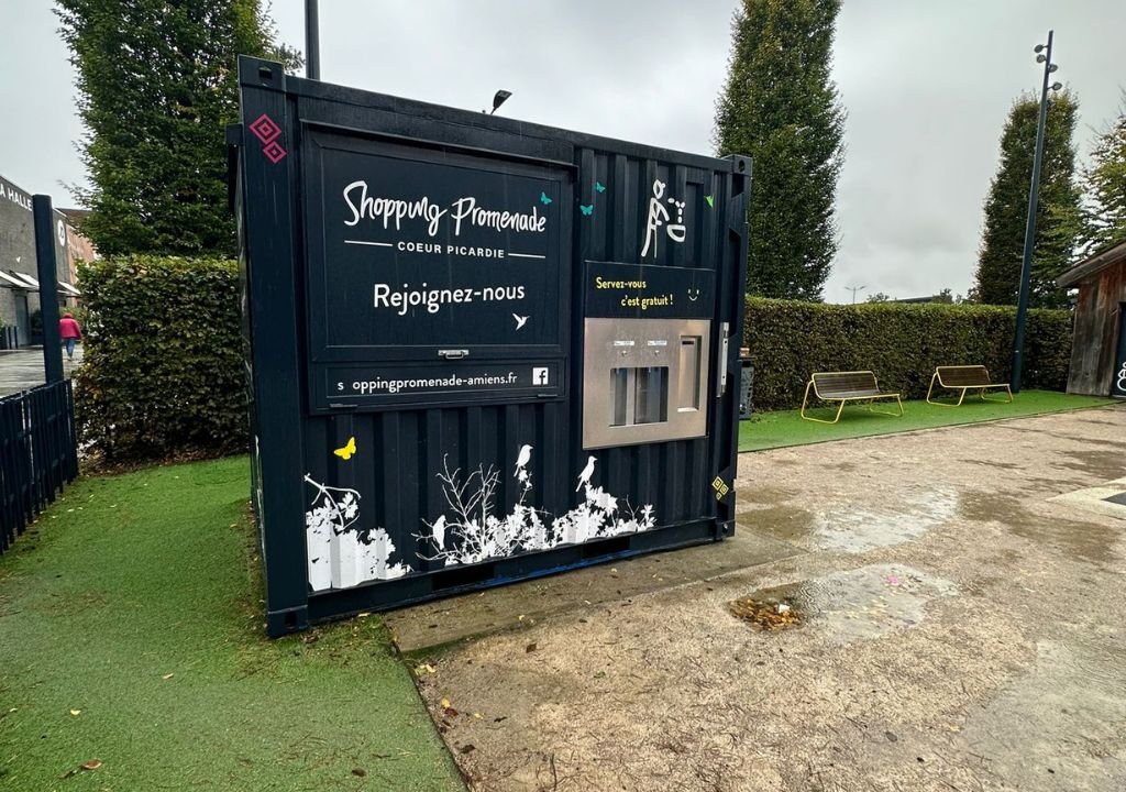 Un conteneur noir portant l'inscription « Shopping Promenade Cœur Picardie », un lien vers un site web et une fontaine à eau avec la mention « Servez-vous, c'est gratuit ! » se trouve dans un espace extérieur agrémenté d'arbres, de bancs et d'un sol humide.