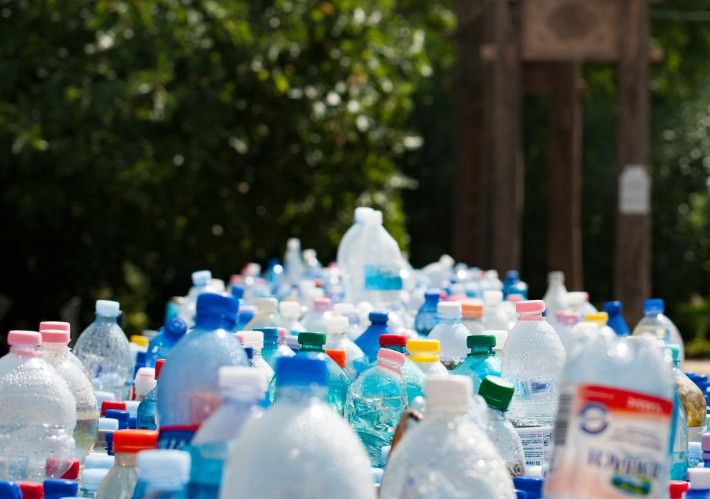 Une grande collection de bouteilles en plastique, principalement des bouteilles d'eau et de boissons, sont regroupées à l'extérieur avec de la verdure et des arbres flous à l'arrière-plan.