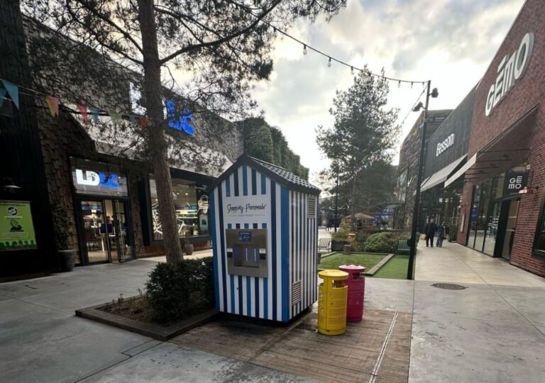 Un petit stand de limonade rayé de bleu et de blanc, surmonté d'une enseigne indiquant « Simply Lemonade », se dresse dans une rue piétonne bordée de boutiques, d'arbres, de guirlandes lumineuses et de bacs de tri sélectif colorés, par une journée nuageuse.