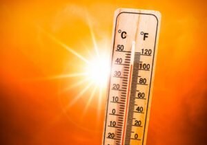 Un thermomètre en bois indique des températures élevées, exprimées à la fois en degrés Celsius et en degrés Fahrenheit, sur fond de ciel orange vif, avec le soleil qui brille intensément à l'arrière-plan.
