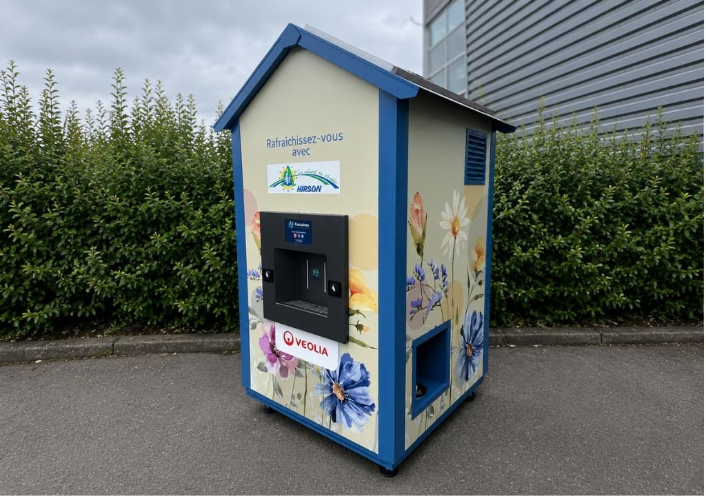 Un distributeur automatique en forme de petite maison, orné de motifs floraux, est installé sur une esplanade pavée, près d'espaces verts et d'un bâtiment. Arborant le logo de Veolia, cette fontaine à eau d'entreprise propose de l'eau fraîche aux passants.