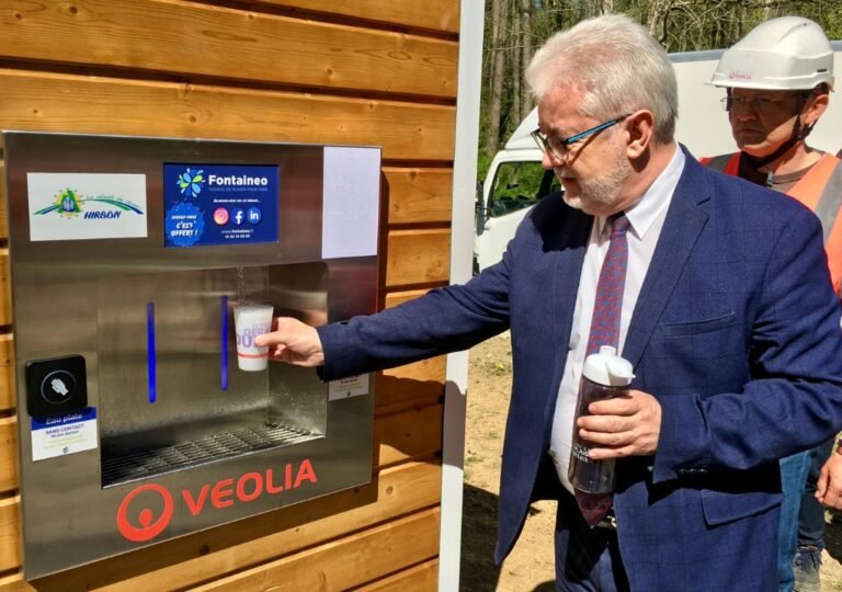 Un homme en costume remplit un verre d'eau à la fontaine à eau d'entreprise VEOLIA, tandis qu'une autre personne, coiffée d'un casque de chantier, se tient à proximité. Le distributeur, fixé à un mur en bois, est orné d'autocollants et de badges.