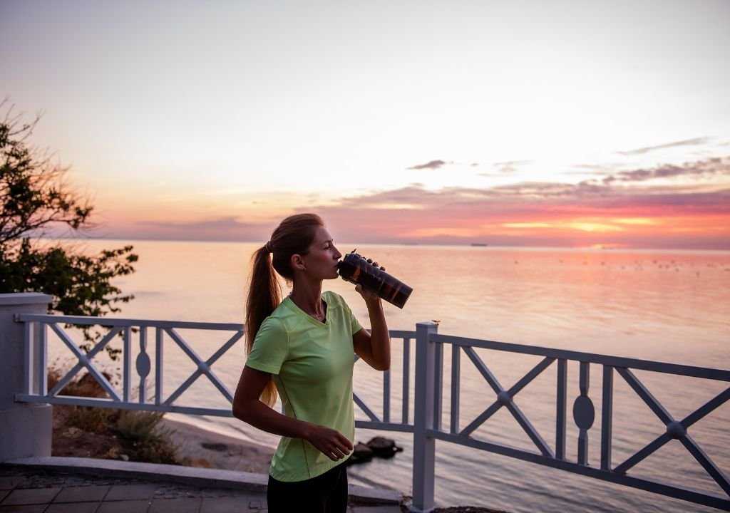 Une femme vêtue d'un t-shirt vert fluo boit à la bouteille, debout près d'une clôture blanche donnant sur une mer calme au coucher du soleil. Le ciel est partiellement nuageux, parsemé de teintes roses et orangées.