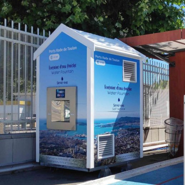 Une grande fontaine à eau, labellisée « Ports Rade de Toulon » et « Fontaine d'eau fraîche », se dresse près d'une zone clôturée. Elle est de couleur bleue et dispose d'un affichage numérique et d'un distributeur d'eau. Une petite poubelle se trouve à proximité.