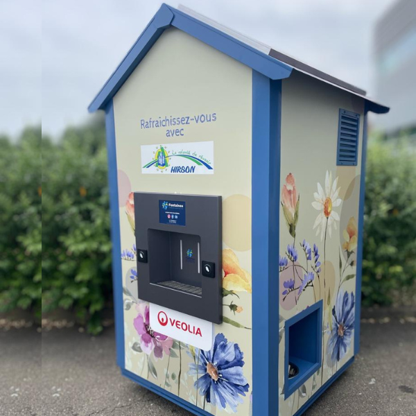 Une fontaine à eau bleue et beige, décorée de fleurs, trône en extérieur. La structure, siglée « Véolia », affiche le texte « Rafraîchissez-vous » et dispose d'un distributeur d'eau sur sa façade. Des buissons et un bâtiment apparaissent en arrière-plan.