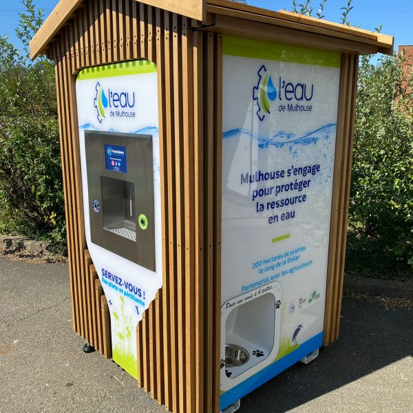 Un kiosque en bois avec distributeur d'eau est installé sur une place pavée. Des panneaux en français, illustrant la politique d'économie d'eau de « l'Eau de Mulhouse », y sont affichés. À proximité, des arbustes sont visibles sous un ciel bleu clair.