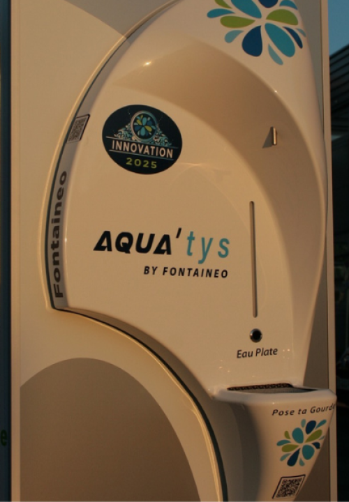 aqua’tys-fontaineo-fontaine-à-eau-réseaux Gros plan sur un distributeur d'eau AQUA'tys de Fontaigueo, au design arrondi, avec l'inscription « Innovation 2025 ». Il est doté de fentes étiquetées « Eau Plate » pour la distribution d'eau plate, ainsi que du logo et de l'adresse du site web.