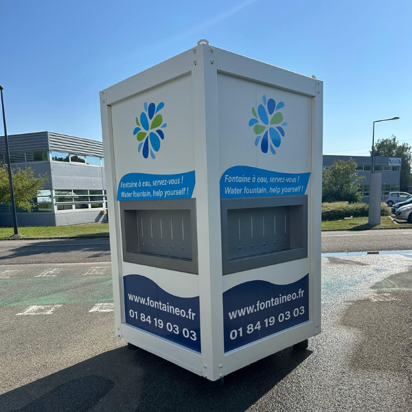 Grande fontaine à eau blanche ornée d'un logo fleuri bleu et vert. « Fontaine à eau, servez-vous ! » imprimé sur les côtés. Coordonnées et URL du site web en bas. Située en extérieur par beau temps.