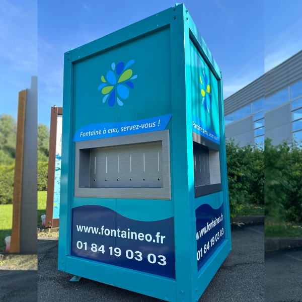 Un grand distributeur d'eau turquoise orné d'un logo floral en haut, d'une zone de distribution au centre et des coordonnées en bas. Il est situé en extérieur, près d'un bâtiment et d'un espace vert.