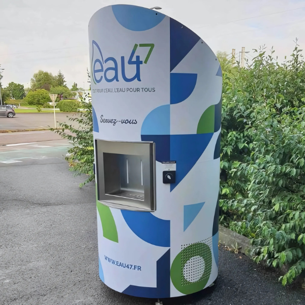 Un distributeur d'eau moderne au design incurvé est installé en extérieur, près d'un arbuste. Il est équipé d'un écran, d'un bouton et de l'inscription « Eau47 » avec un lien vers son site web. Le quartier est bordé d'asphalte et de verdure.