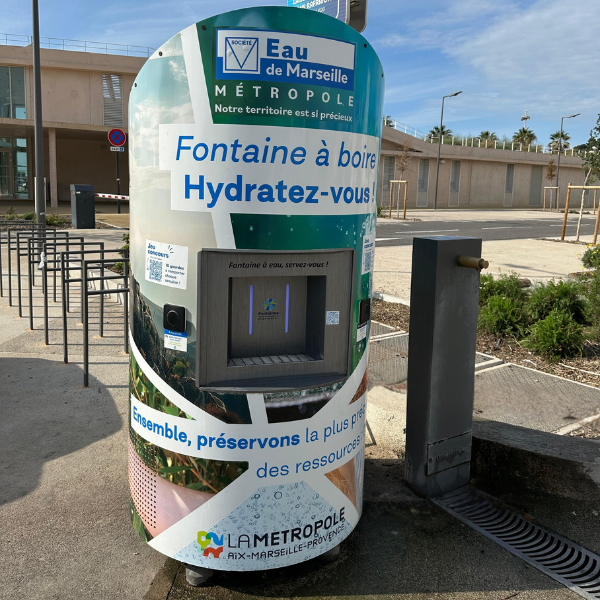 À Marseille, une fontaine cylindrique propose de l'eau potable avec un message invitant à rester hydraté. L'extérieur est décoré d'images et de logos sur le thème de l'eau. Un écran numérique et un distributeur sont visibles. Les bâtiments et les plantes environnants apparaissent en arrière-plan.