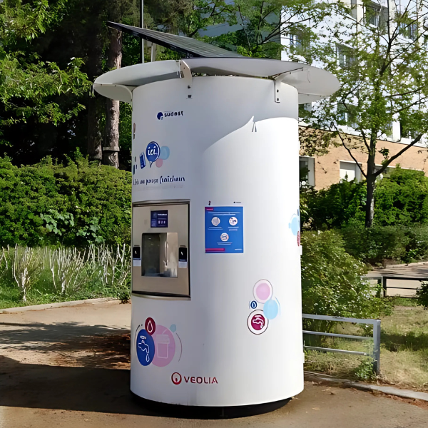 Un distributeur automatique de boissons cylindrique blanc est installé en extérieur, surmonté d'un panneau solaire. La surface est ornée de logos, d'un écran et d'un espace de distribution. Entouré de verdure et d'allées, un bâtiment est visible en arrière-plan.