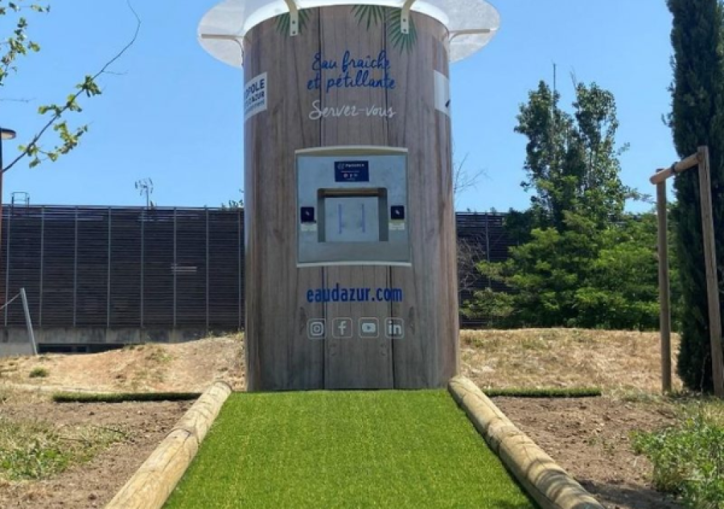 Un distributeur d'eau se dresse sur une pelouse artificielle, encadré par deux poutres en bois. Il affiche l'inscription « eau fraîche et pétillante » et des liens vers les réseaux sociaux. Des arbres et une clôture apparaissent en arrière-plan, sous un ciel dégagé.