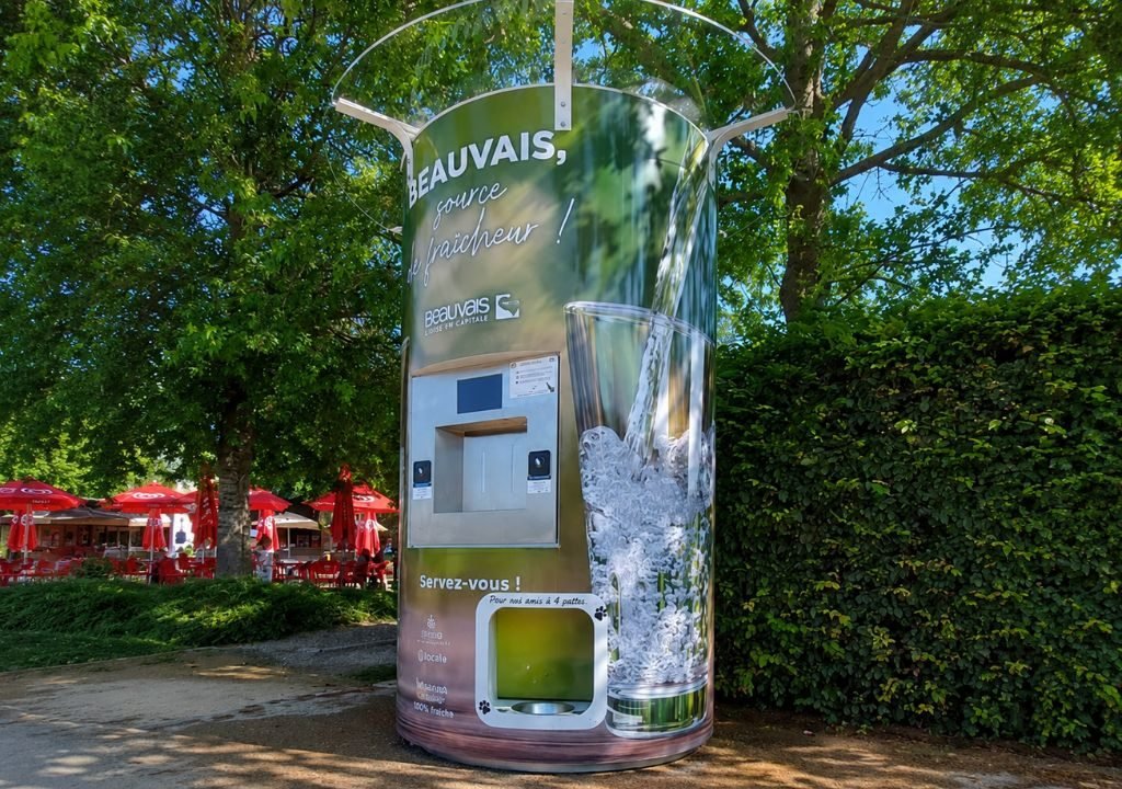 Un grand distributeur d'eau en plein air, sur lequel figure l'image d'un verre d'eau, se trouve sous les arbres, à côté d'un sentier. On aperçoit des parasols rouges et des tables à l'arrière-plan, à gauche.