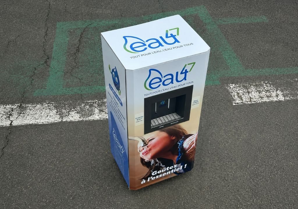 Un distributeur d'eau portant la marque « eau4 » est installé sur le trottoir. Sur le boîtier figurent la photo d'un enfant en train de boire de l'eau, un texte en français et des instructions expliquant comment se servir de l'eau.