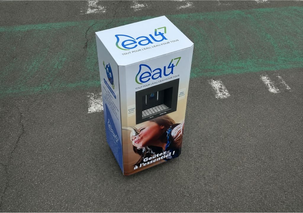 Une boîte portant la marque « eau4 » et illustrée d'un distributeur d'eau et d'un enfant en train de boire est posée sur une surface asphaltée où se dessinent des lignes peintes en blanc et vert, désormais effacées.