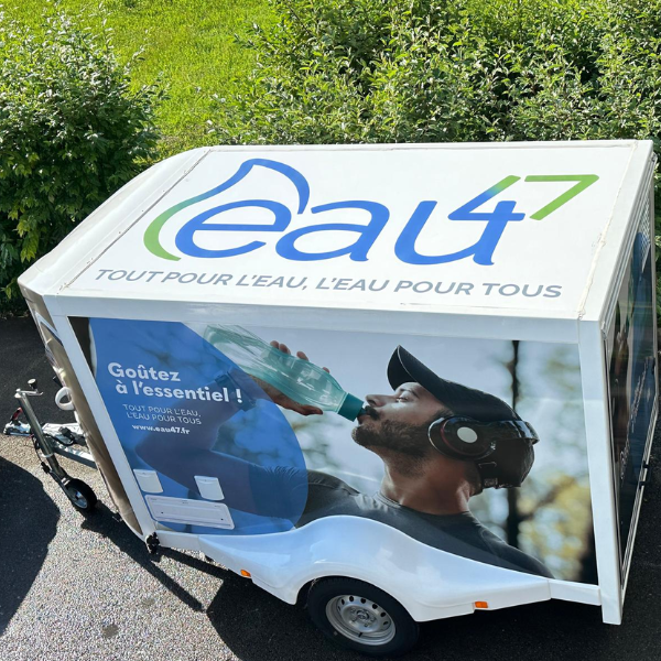 Une remorque blanche portant le logo « eau47 » et l'inscription « Tout pour l'eau, l'eau pour tous » est garée sur une route. On y voit un homme portant une casquette et un casque, buvant de l'eau à la bouteille. Un feuillage vert apparaît en arrière-plan.