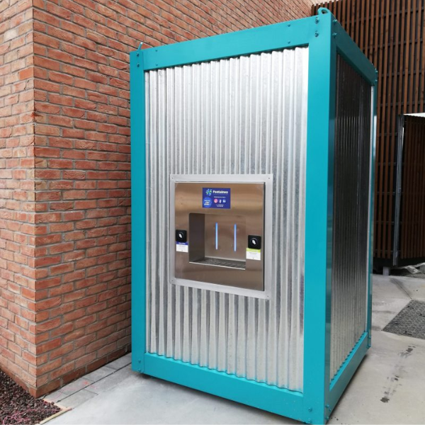 Un kiosque métallique rectangulaire avec un cadre bleu sarcelle et des panneaux argentés ondulés, doté d'un écran central et d'une fente de distribution, est positionné sur une zone pavée à côté d'un mur de briques.