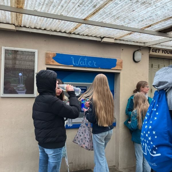 Plusieurs personnes, principalement des filles, se tiennent sous un abri près d'une fontaine murale étiquetée « Eau ». Une fille boit à la bouteille tandis que d'autres attendent ou discutent. Le temps semble pluvieux ou humide.