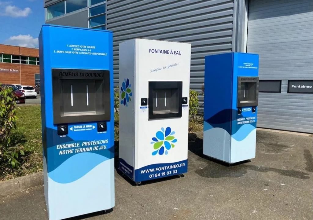 Trois bornes de remplissage d'eau en extérieur sont installées sur une surface pavée devant un bâtiment. Les bornes sont bleues et blanches ; elles comportent des instructions et un logo sur le thème de l'eau. Une ombre se dessine sur le sol.