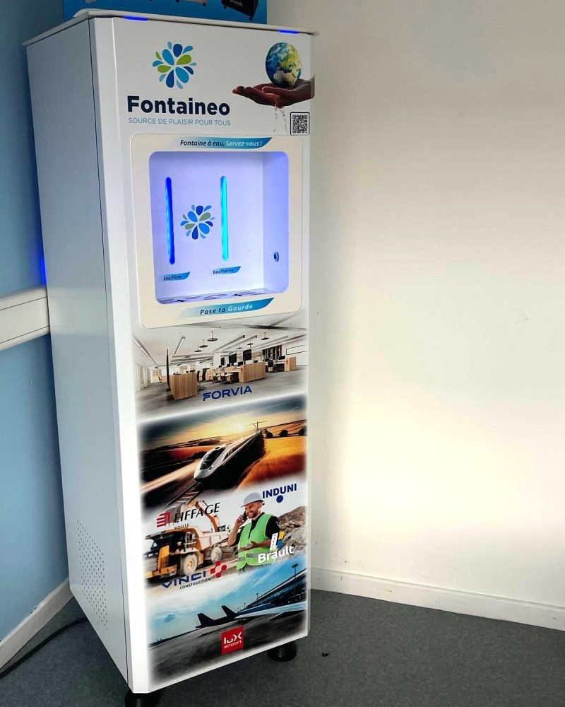 fontaine-à-eau-réseau-light Un distributeur automatique blanc portant l'inscription « Fontaineo » est placé contre un mur. La machine est dotée d'un écran numérique en haut et de diverses publicités en bas. Elle repose sur quatre petites roues sur un sol gris.
