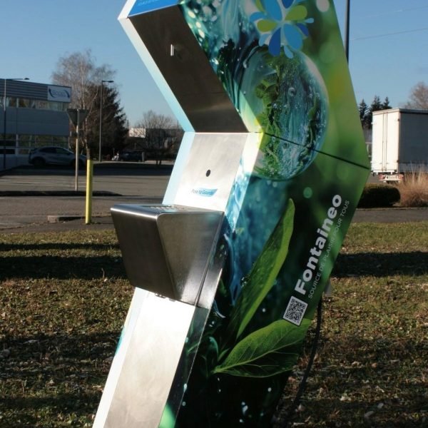 Un grand distributeur d'eau étiqueté « Fontaineo » se trouve à l'extérieur sur un terrain herbeux. La machine présente un design sur le thème de la nature avec des images de feuilles et de gouttes d'eau. Elle se trouve sous un ciel clair, avec quelques arbres et bâtiments en arrière-plan.