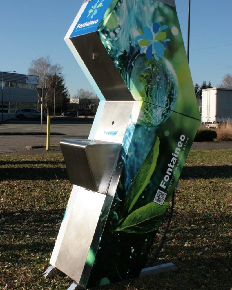fontaine-à-eau-réseau-nature Un grand distributeur d'eau étiqueté « Fontaineo » se trouve à l'extérieur sur un terrain herbeux. La machine présente un design sur le thème de la nature avec des images de feuilles et de gouttes d'eau. Elle se trouve sous un ciel clair, avec quelques arbres et bâtiments en arrière-plan.