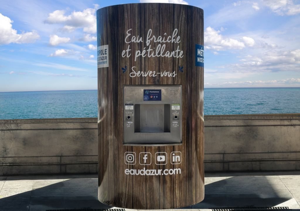 fontaine-a-eau-réseau-port Un distributeur d'eau public avec des panneaux en bois se trouve sur une promenade en bord de mer. L'enseigne indique "Eau fraîche et pétillante Servez-vous", offrant de l'eau fraîche et pétillante. Des icônes de médias sociaux et un site web sont visibles.
