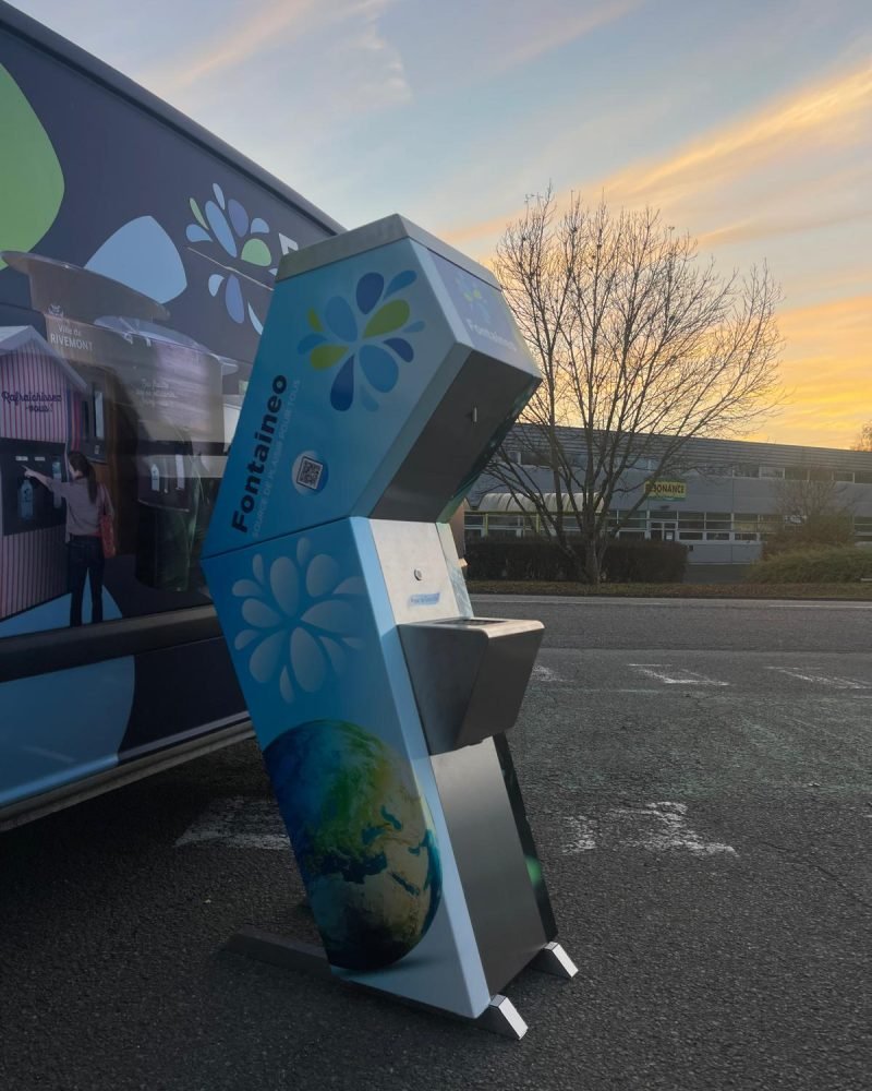 fontaine-à-eau-réseau-primo Un kiosque de distribution d'eau autonome avec la marque « Fontaineo » est positionné sur une zone pavée. Il présente des graphismes floraux bleus et une image de la terre. Un arbre et un bâtiment sont visibles en arrière-plan sous un ciel aux nuages vaporeux.
