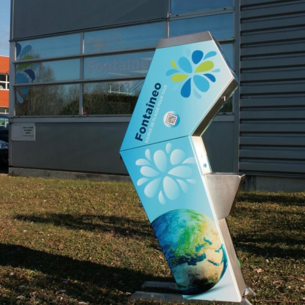 Un distributeur d'eau baptisé « Fontaineo » est installé à l'extérieur. La station est décorée de bleu et de blanc avec des motifs de gouttes d'eau et une image de la Terre. Elle se trouve sur une zone herbeuse près d'un bâtiment moderne aux fenêtres réfléchissantes.