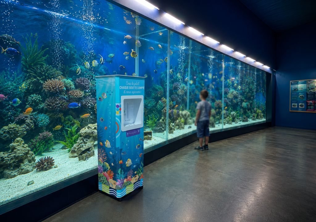 Un jeune garçon se tient devant un grand aquarium rempli de poissons colorés et de coraux. La pièce a des murs bleus, des sols polis et, près du kiosque d'information, une fontaine à eau parc d'attraction où les visiteurs peuvent se rafraîchir.