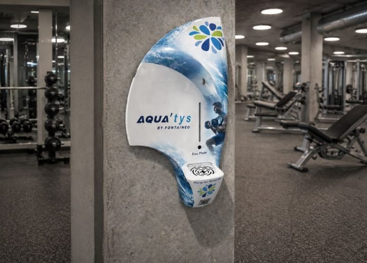 Un distributeur d'eau mural portant la mention « AQUA'tys by Fontaigne », sur lequel figure l'image d'une personne en train de nager, est fixé à un pilier en béton dans une salle de sport, avec divers appareils de musculation en arrière-plan.