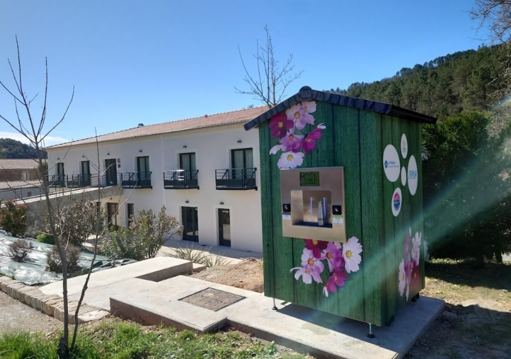 Un petit kiosque vert avec des motifs de fleurs roses se trouve sur une plate-forme en béton près d'un bâtiment de deux étages avec des balcons et entouré d'arbres sous un ciel bleu clair.