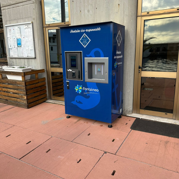 Un distributeur d'eau bleu, labélisé « Fontaine éco-responsable », se trouve à l'extérieur d'un bâtiment. À côté d'un panneau d'affichage et de portes vitrées, il est installé sur un sol carrelé rouge, contre un mur en béton.
