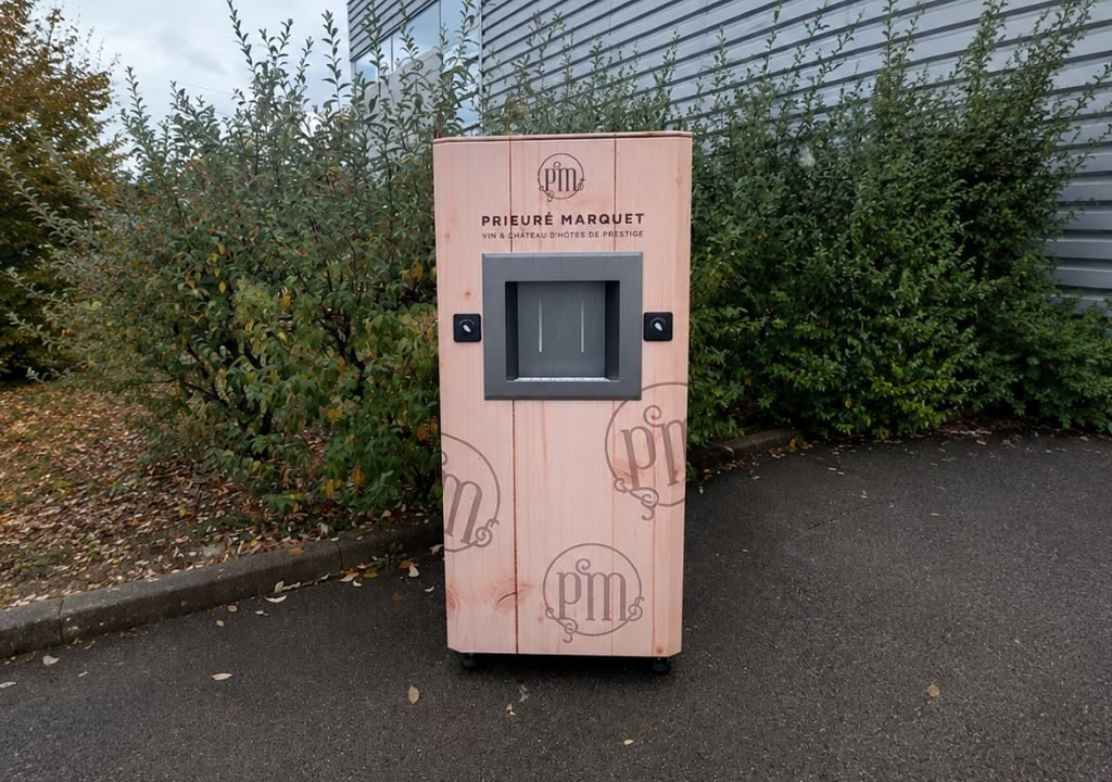 Un distributeur automatique en bois portant l'inscription « Prieuré Marquet » se trouve sur une esplanade pavée, près de buissons et d'un bâtiment métallique. L'appareil est doté d'un écran central et affiche une adresse de site web ainsi qu'un logo sur sa face avant.