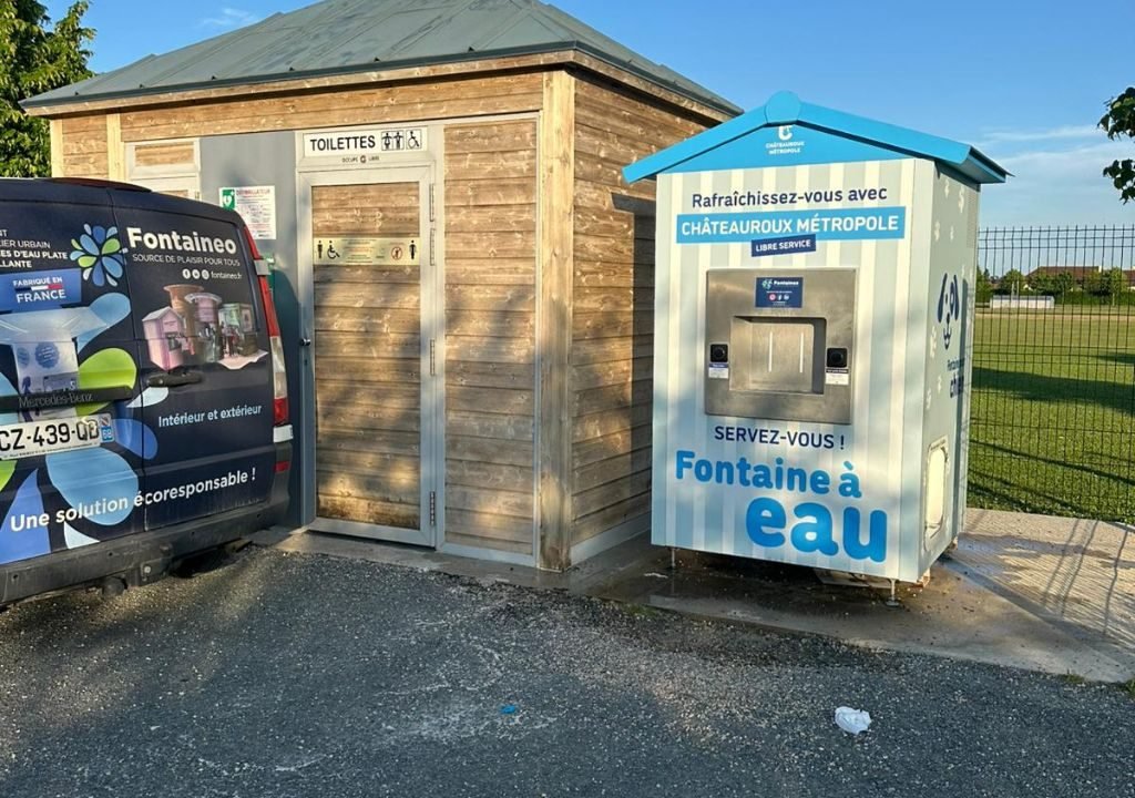 Une camionnette grise et un distributeur d'eau sont garés à côté de toilettes publiques en bois portant l'inscription « Toilettes », dans un cadre en plein air baigné de soleil. On aperçoit de l'herbe, une clôture et des arbres à l'arrière-plan.
