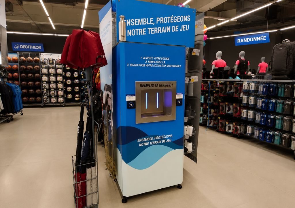 Une borne de remplissage d'eau située à l'intérieur d'un magasin d'articles de sport, accompagnée d'un texte en français incitant à l'éco-responsabilité. La borne, de couleur bleue et blanche, est installée parmi des rayons proposant des sacs à dos, des vêtements et des équipements sportifs, sous un éclairage vif.