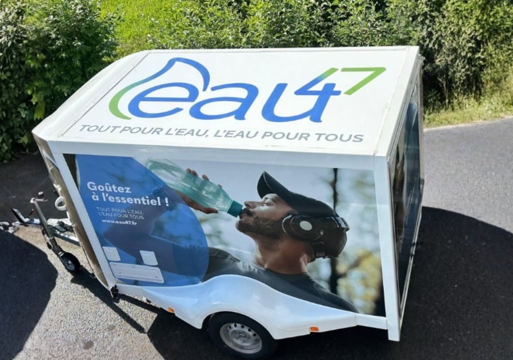 Une remorque blanche portant les inscriptions « eau47 » et « Tout pour l'eau, l'eau pour tous » sur le toit. Sur le côté, on voit un homme en train de boire à une bouteille. La remorque est garée sur une route, à côté d'un espace vert.