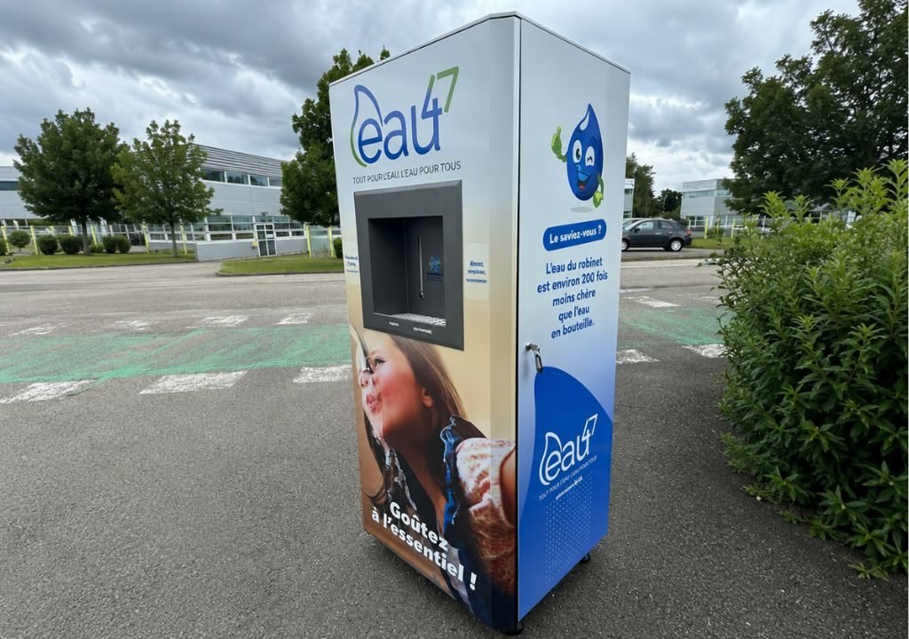 Une fontaine à eau publique d'entreprise portant le nom « Eau4 » se trouve sur un parking ; elle est ornée d'une photo représentant une fillette en train de boire de l'eau et d'un texte en français décrivant sa mission : fournir de l'eau potable à tous. Des arbres et des bâtiments apparaissent à l'arrière-plan.
