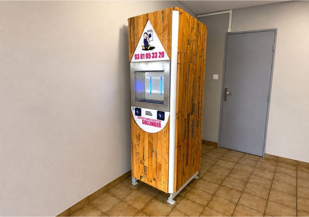 Un distributeur automatique, dont la façade est recouverte de boiseries, se trouve dans un coin d'une pièce carrelée, près d'une porte grise fermée, à côté d'une fontaine à eau d'entreprise. Sur sa façade, on peut lire un numéro de téléphone et le nom « SOLLINGER ».