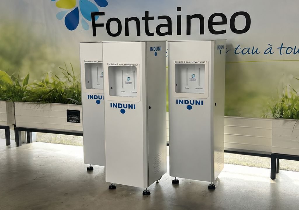 Trois distributeurs d'eau blanche portant la marque « INDUNI » sont alignés à l'intérieur, devant un mur orné d'un grand logo « Fontaineo » et de plantes décoratives.
