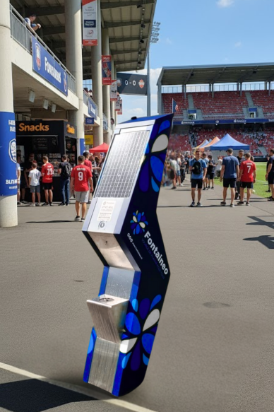 fontaine-primo-fontaine-a-eau-reseau Une fontaine d'eau publique alimentée par l'énergie solaire et portant l'étiquette "Fontaineo" se trouve à l'extérieur d'un stade de sport. Des personnes en maillot marchent à proximité et des spectateurs sont visibles dans les gradins.