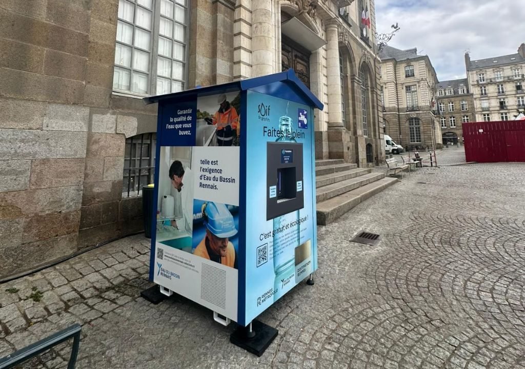 Un petit kiosque bleu et blanc se dresse dans une rue pavée, près d'un bâtiment en pierre à colonnes. Le kiosque présente des images et des textes sur la qualité de l'eau et est équipé d'un écran numérique et d'un distributeur.