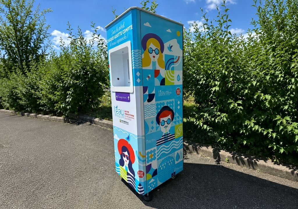 Un distributeur automatique coloré est installé sur le trottoir, près de buissons verts. Il est orné d'illustrations représentant des personnes portant des lunettes de soleil, de motifs aquatiques et de formes aux couleurs vives, sous un ciel partiellement nuageux.