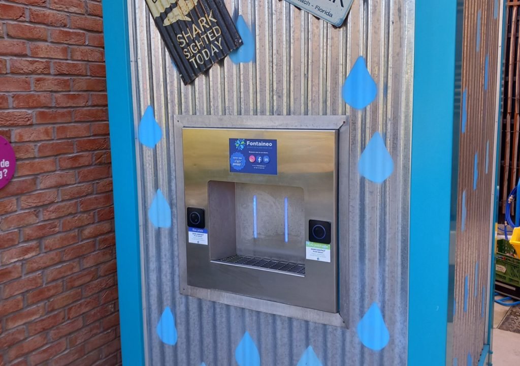 fontaine strasbourg Une station de remplissage d'eau en métal avec un cadre turquoise, des gouttes d'eau peintes en bleu et deux panneaux inclinés au-dessus indiquant « DANGER REQUIN » et « SURF À VOS RISQUES ET PÉRILS REQUIN APERÇU AUJOURD'HUI », contre un mur de briques.
