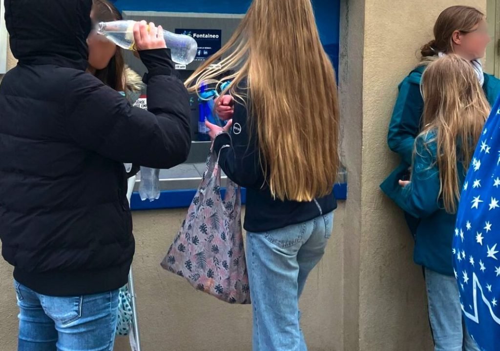 Quatre jeunes se tiennent devant une station de remplissage d'eau ; l'un boit dans une bouteille, l'autre tient un sac réutilisable et l'un d'eux a des béquilles. Tous sont habillés de manière décontractée, avec des vestes et des jeans.