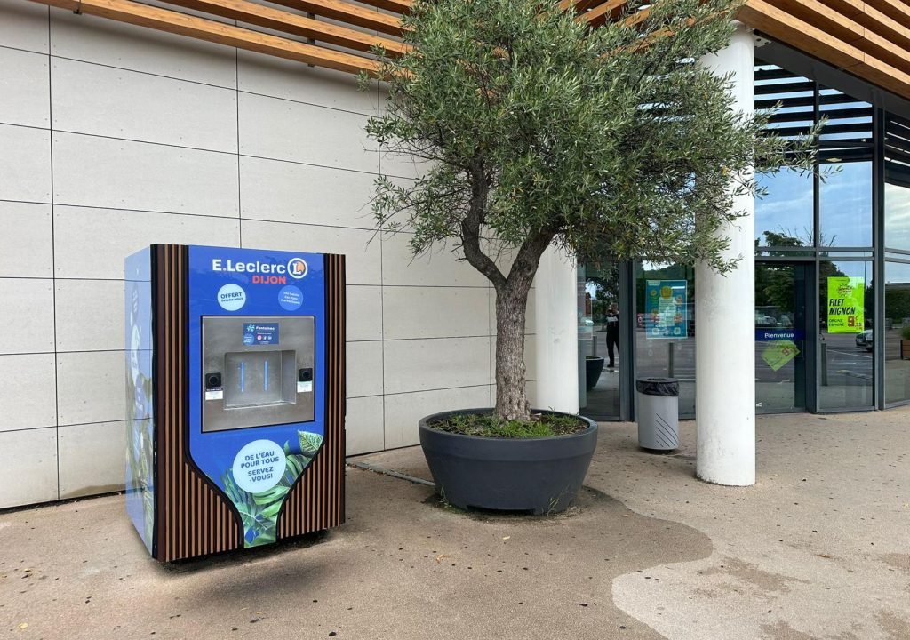 Une fontaine à eau portant l'inscription « E.Leclerc Dijon » se trouve à côté d'un grand arbre en pot, devant un bâtiment doté d'un auvent moderne et d'une entrée vitrée. Une poubelle se trouve à proximité.