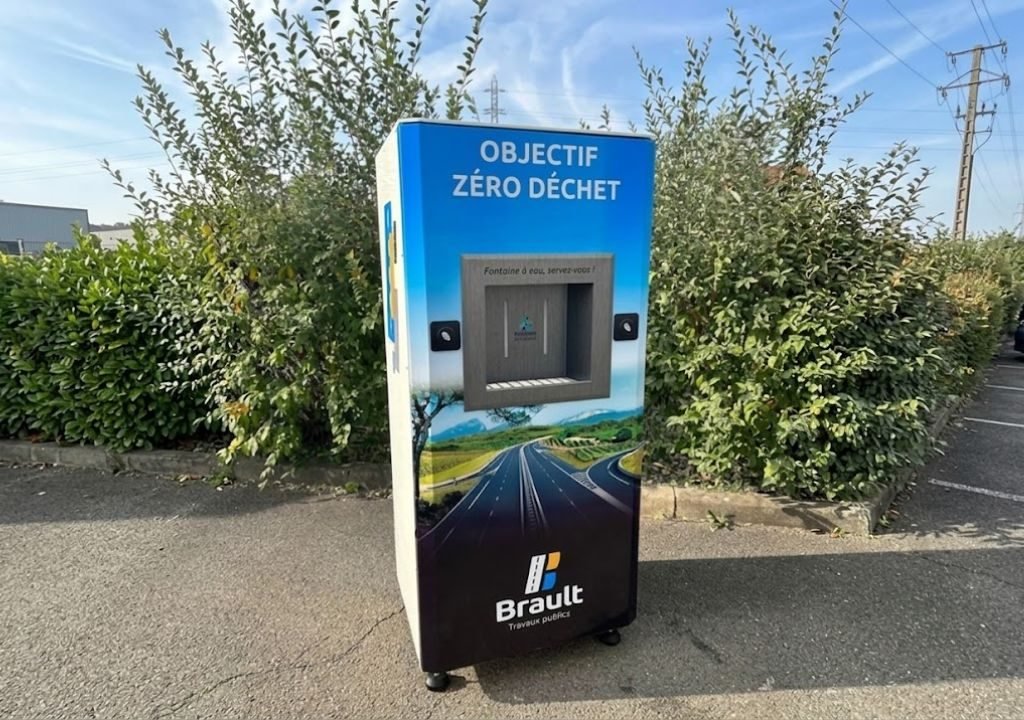 Une machine de recyclage de type distributeur automatique, portant l'inscription « Objectif Zéro Déchet », est installée à l'extérieur sur le trottoir, près de buissons, à côté d'une fontaine à eau de chantier. La machine arbore le logo Brault ainsi qu'une illustration représentant une route sous un ciel bleu parsemé de nuages.