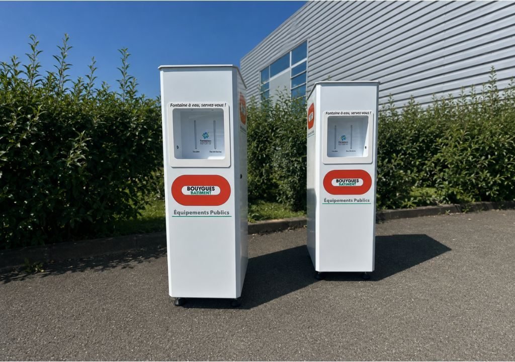 Deux fontaines à eau de chantier blanches, munies d'étiquettes rouges et vertes, sont installées sur une aire pavée en plein air, à côté de buissons verts et d'un bâtiment métallique doté de grandes fenêtres.