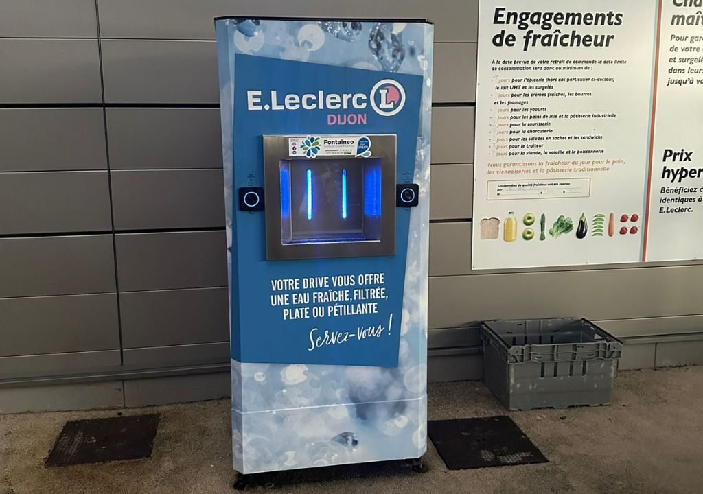 Un distributeur d'eau E.Leclerc bleu est adossé à un mur gris et propose de l'eau fraîche, filtrée, plate ou gazeuse. Il est doté de zones de distribution éclairées et d'un texte promotionnel en français sur la face avant.